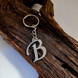 May include: A silver-colored metal keychain featuring the letter "B". The letter is stylized with a decorative font and is attached to a chain and a keyring. The keychain is displayed on a piece of dark brown wood.