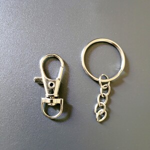 May include: A silver-colored keychain hardware set, including a lobster clasp and a split ring with a chain. The clasp has a lever for easy attachment. The set is designed for creating custom keychains or attaching items to bags.