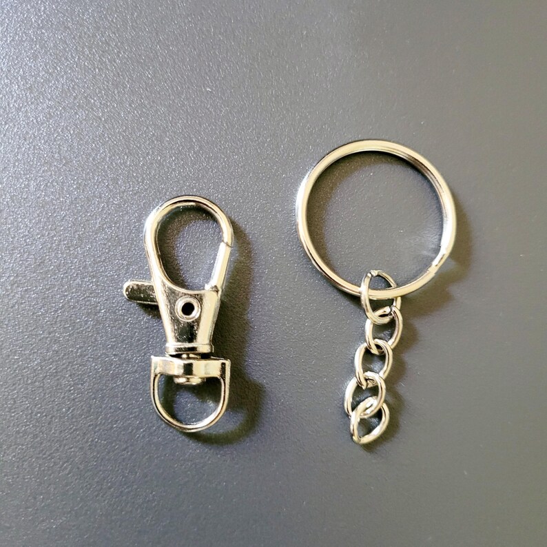 May include: A silver-colored keychain hardware set, including a lobster clasp and a split ring with a chain. The clasp has a lever for easy attachment. The set is designed for creating custom keychains or attaching items to bags.
