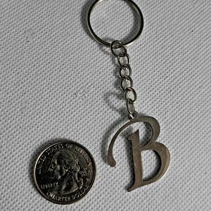 May include: Silver-colored metal keychain with the letter "B" pendant. The keychain includes a ring and a chain of small links. A U.S. quarter coin is next to the keychain for size comparison.
