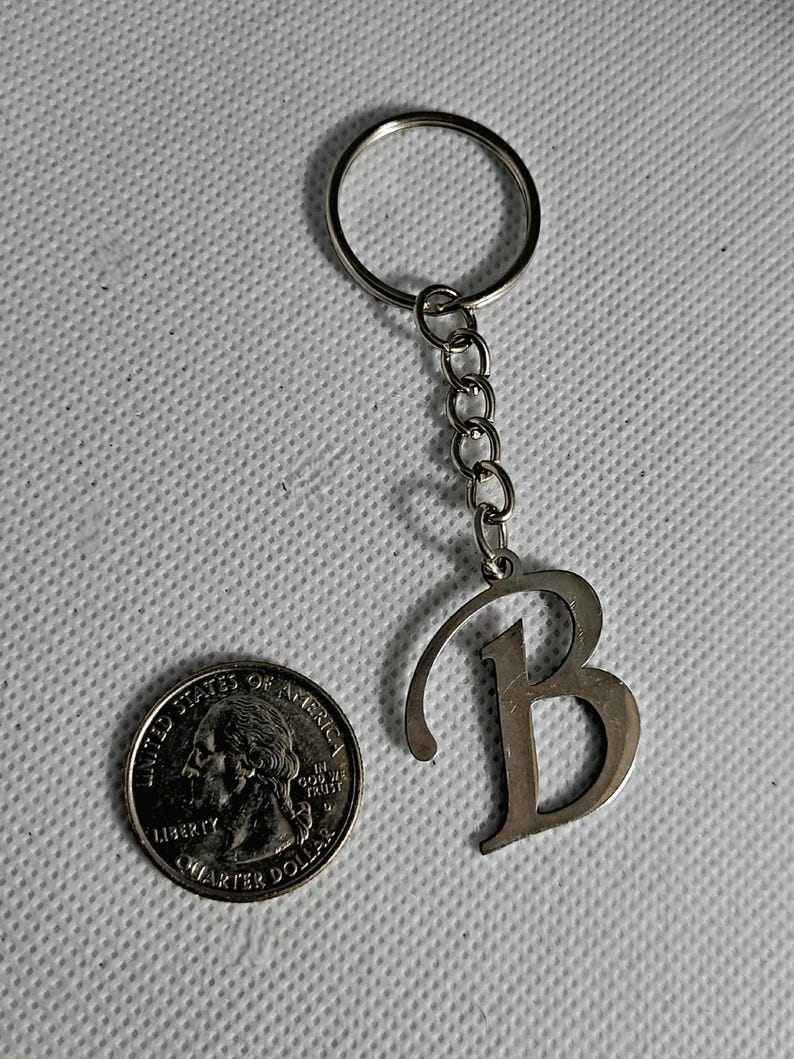 May include: Silver-colored metal keychain with the letter "B" pendant. The keychain includes a ring and a chain of small links. A U.S. quarter coin is next to the keychain for size comparison.