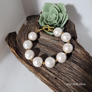 May include: A bracelet featuring large, round, white faux pearls alternating with small, metallic gold-toned beads. The piece is secured with a gold-toned toggle clasp. The text "Mon Petit Bijou" is visible in the bottom right corner.