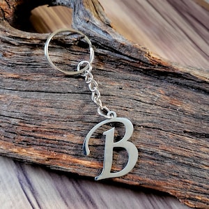 May include: A silver-colored metal keychain featuring the letter "B" in a cursive font. The keychain includes a chain and a ring for attaching keys. The background is a piece of weathered wood.
