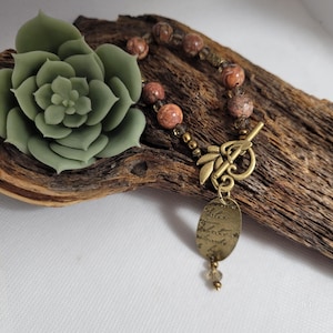May include: A handmade bracelet with reddish-brown beads and a decorative clasp. The bracelet features a brass oval charm with etched text and a small crystal pendant. A green succulent and a piece of driftwood are also in the image.