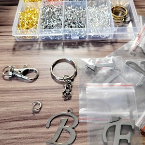 May include: A collection of jewelry-making supplies, including gold, silver, and copper-colored jump rings, keychains, and letter charms. The items are organized in a clear plastic container and small plastic bags.