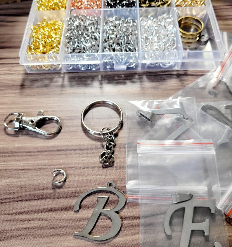 May include: A collection of jewelry-making supplies, including gold, silver, and copper-colored jump rings, keychains, and letter charms. The items are organized in a clear plastic container and small plastic bags.