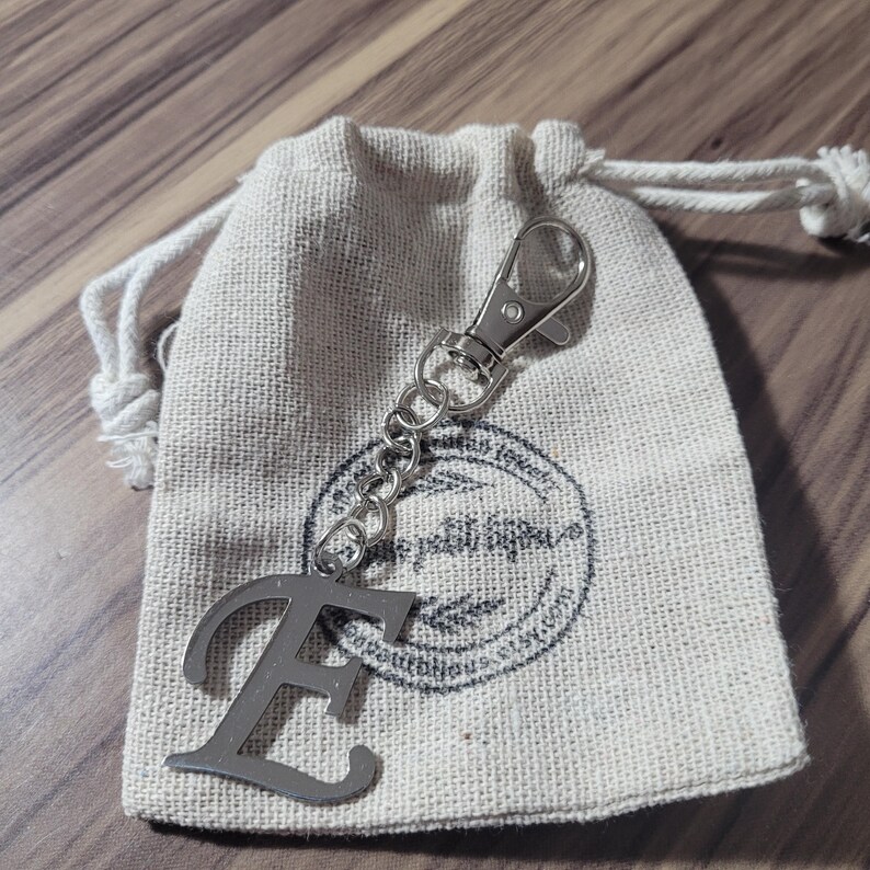 May include: A silver metal keychain in the shape of the letter "E" with a chain and clasp, resting on a small beige drawstring bag. The bag has a printed logo. The background is a wooden surface.
