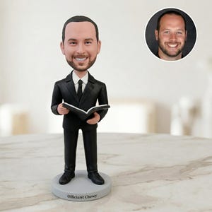 May include: A bobblehead figurine of a man in a black suit, holding an open book. The figurine has a smiling face and is standing on a gray base with the text "Officiant Chewy". A smaller image of the man's face is in the background.