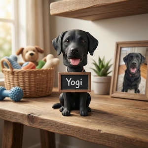 May include: A black Labrador statue with a small chalkboard sign that reads "Yogi" around its neck. A framed photo of a black Labrador is on the right. A basket of dog toys and a dumbbell are in the background.