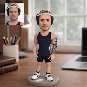 May include: A personalized bobblehead figurine of a man in a navy blue wrestling singlet and black shorts, wearing a head guard. The figure has a detailed tattoo on one arm and is standing on a gray base, wearing white sneakers.