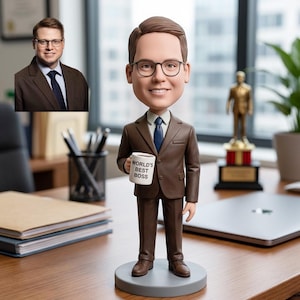 Op de afbeelding: Een bobblehead-beeldje van een man in een bruin pak, met een mok met de tekst "WORLD'S BEST BOSS". Het beeldje staat op een grijze basis. Een kleinere afbeelding van de man in pak en bril staat op de achtergrond.
