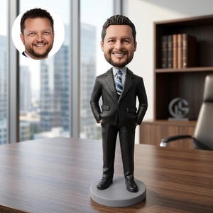 May include: A bobblehead figurine of a man in a black suit, a white and blue striped tie, and black shoes. The figurine stands on a grey circular base. A smaller image of the man's face is in the upper left corner.