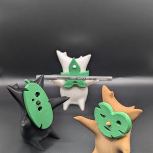Peut inclure: Trois petites figurines imprimées en 3D, chacune avec une couleur différente et un masque en forme de feuille verte. Les figurines sont debout sur une surface noire. La figurine blanche tient un stylo avec le texte "Round Stic - M".