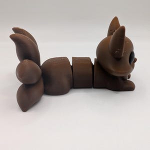 Pu&ograve; includere: Una statuetta articolata e segmentata di colore marrone che raffigura una creatura stilizzata con grandi orecchie a punta, una testa arrotondata e una coda composta da pi&ugrave; parti. L'oggetto presenta linee di stampa 3D orizzontali.
