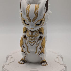 Pu&ograve; includere: Una statuetta stampata in 3D di una creatura stilizzata con segni bianchi e dorati simili a un'armatura. La figura presenta orecchie a punta, grandi occhi chiusi e una superficie strutturata su una base esagonale bianca.