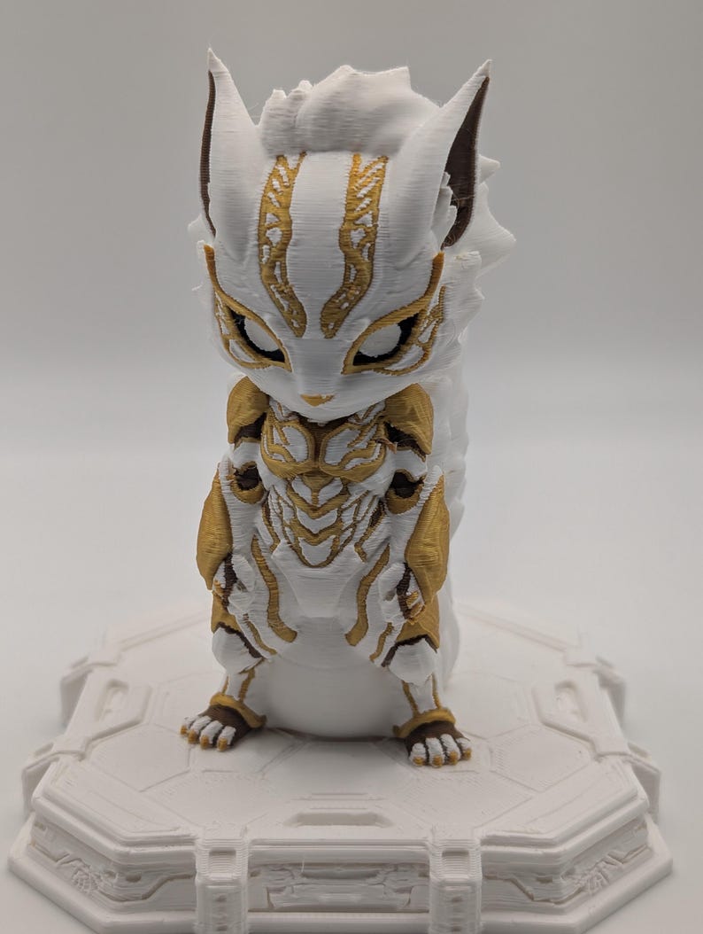 Pu&ograve; includere: Una statuetta stampata in 3D di una creatura stilizzata con segni bianchi e dorati simili a un'armatura. La figura presenta orecchie a punta, grandi occhi chiusi e una superficie strutturata su una base esagonale bianca.