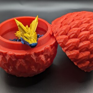 Könnte beinhalten: Eine rote Dracheneier-förmige Schmuckschatulle mit einem goldenen und blauen Origami-Drachen im Inneren.