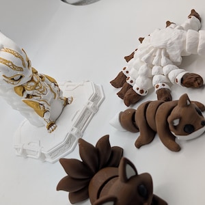 Pu&ograve; includere: Una collezione di quattro statuette articolate che include una creatura bianca e dorata su base, una grande volpe bianca con molte code, una piccola volpe marrone con la punta della coda bianca e una statuetta di volpe marrone con molte code.