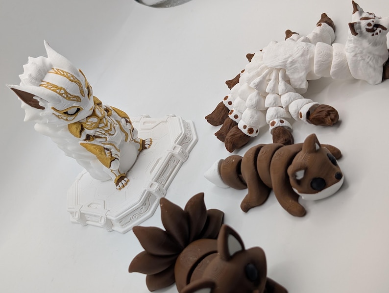 Pu&ograve; includere: Una collezione di quattro statuette articolate che include una creatura bianca e dorata su base, una grande volpe bianca con molte code, una piccola volpe marrone con la punta della coda bianca e una statuetta di volpe marrone con molte code.