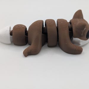 Pu&ograve; includere: Una statuetta di volpe articolata marrone e bianca con corpo e coda segmentati. La volpe ha una testa marrone con occhi neri, accenti bianchi sul muso e una punta bianca sulla coda. Le linee di stampa 3D sono visibili sulla superficie.