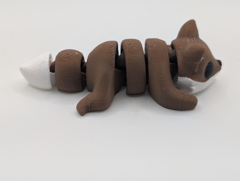 Pu&ograve; includere: Una statuetta di volpe articolata marrone e bianca con corpo e coda segmentati. La volpe ha una testa marrone con occhi neri, accenti bianchi sul muso e una punta bianca sulla coda. Le linee di stampa 3D sono visibili sulla superficie.