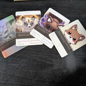 Pu&ograve; includere: Quattro carte da gioco rettangolari con creature fantastiche illustrate. Le carte mostrano personaggi chiamati Delta, Nine-Tailed Spiritfox, The Guardian e Spirit Pup, ciascuna con testo stampato, linee statistiche e illustrazioni uniche.