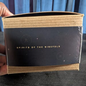 Pu&ograve; includere: Una scatola di cartone rettangolare con una fascia di carta nera avvolta attorno al centro. La fascia mostra il testo dorato "SPIRITS OF THE NINEFOLD" insieme a due timbri circolari contenenti i caratteri cinesi "九" e "尾".