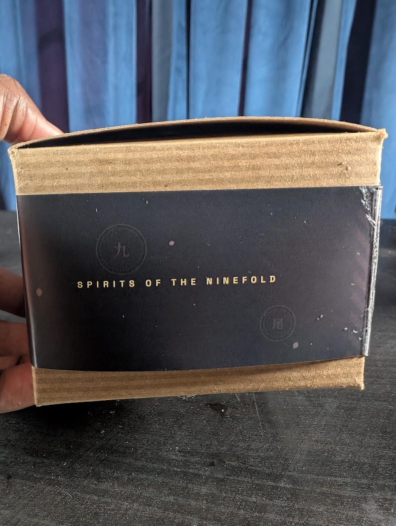 Pu&ograve; includere: Una scatola di cartone rettangolare con una fascia di carta nera avvolta attorno al centro. La fascia mostra il testo dorato "SPIRITS OF THE NINEFOLD" insieme a due timbri circolari contenenti i caratteri cinesi "九" e "尾".