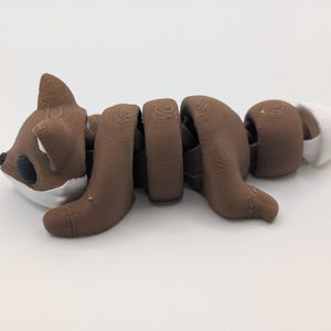 Pu&ograve; includere: Una statuetta di volpe articolata marrone e bianca con una texture stampata in 3D a strati. La volpe presenta una testa arrotondata con occhi neri, orecchie a punta e un corpo segmentato che consente il movimento, terminando con una coda bianca.