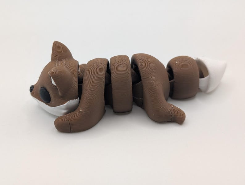 Pu&ograve; includere: Una statuetta di volpe articolata marrone e bianca con una texture stampata in 3D a strati. La volpe presenta una testa arrotondata con occhi neri, orecchie a punta e un corpo segmentato che consente il movimento, terminando con una coda bianca.
