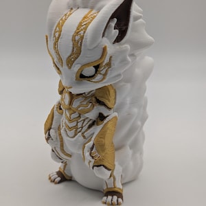 Pu&ograve; includere: Una statuetta stampata in 3D di una creatura stilizzata nei colori bianco e oro. La figura presenta una testa simile a quella di una volpe con orecchie a punta, intricati segni dorati sul viso e sul busto e una grande coda bianca.