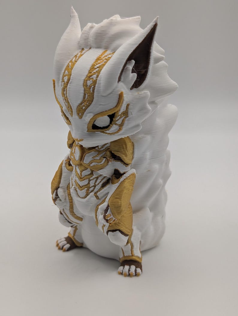 Pu&ograve; includere: Una statuetta stampata in 3D di una creatura stilizzata nei colori bianco e oro. La figura presenta una testa simile a quella di una volpe con orecchie a punta, intricati segni dorati sul viso e sul busto e una grande coda bianca.