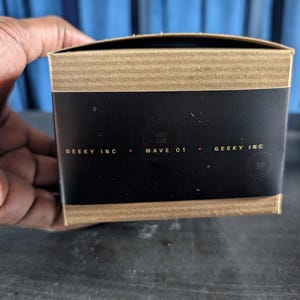 Pu&ograve; includere: Una scatola di cartone rettangolare con una fascia centrale nera che riporta il testo GEEKY INC e WAVE 01 in lettere dorate. I bordi superiore e inferiore della scatola sono in cartone ondulato marrone e sono visibili due loghi circolari.