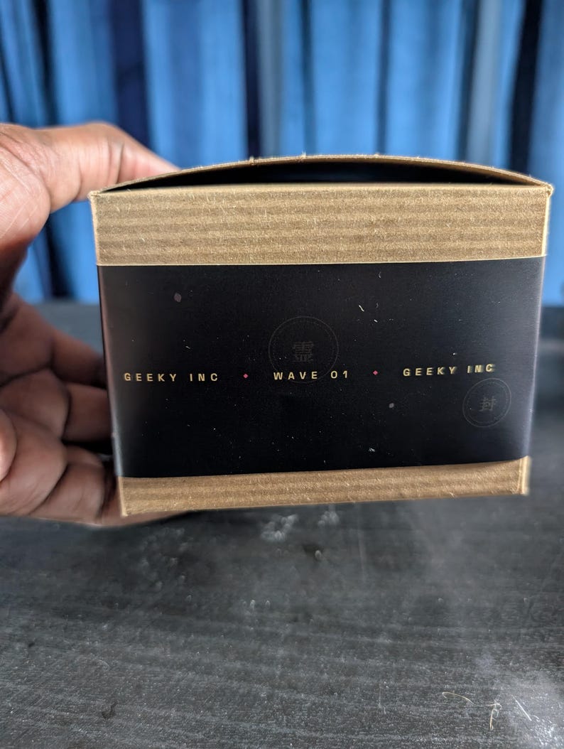 Pu&ograve; includere: Una scatola di cartone rettangolare con una fascia centrale nera che riporta il testo GEEKY INC e WAVE 01 in lettere dorate. I bordi superiore e inferiore della scatola sono in cartone ondulato marrone e sono visibili due loghi circolari.
