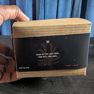 Pu&ograve; includere: Una scatola rettangolare di cartone avvolta da una fascia di carta nera. La fascia riporta il testo "FOUR IN THE LOOT POOL. ONE PULL PER SEAL.", "SHIRO 1 IN 10", "GEEKYINC.COM" e i caratteri giapponesi "封", "狐" e "神" con un grafico di una coda di volpe.