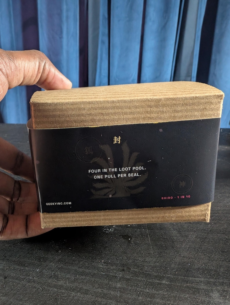Pu&ograve; includere: Una scatola rettangolare di cartone avvolta da una fascia di carta nera. La fascia riporta il testo "FOUR IN THE LOOT POOL. ONE PULL PER SEAL.", "SHIRO 1 IN 10", "GEEKYINC.COM" e i caratteri giapponesi "封", "狐" e "神" con un grafico di una coda di volpe.