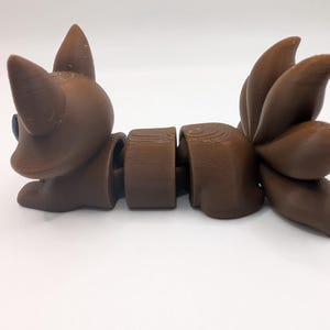 Pu&ograve; includere: Una statuetta di volpe articolata marrone con un corpo segmentato e una grande coda folta composta da tre sezioni distinte. La figura presenta orecchie a punta, una superficie liscia con linee di stampa 3D visibili e un occhio scuro.