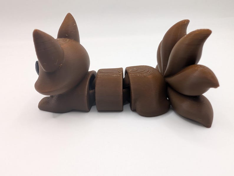 Pu&ograve; includere: Una statuetta di volpe articolata marrone con un corpo segmentato e una grande coda folta composta da tre sezioni distinte. La figura presenta orecchie a punta, una superficie liscia con linee di stampa 3D visibili e un occhio scuro.