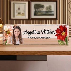 May include: A clear acrylic desk nameplate featuring a woman's photo, the name "Angelina Miller", and the title "FINANCE MANAGER". The nameplate is decorated with floral designs in shades of red and peach.