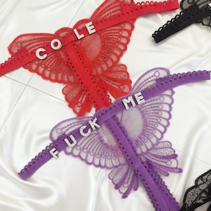May include: Red, purple, and black lace lingerie with rhinestone lettering spelling out "COLE" and "FUCK ME". The lingerie features intricate floral patterns and a delicate, see-through design. The background is a white, silky fabric.