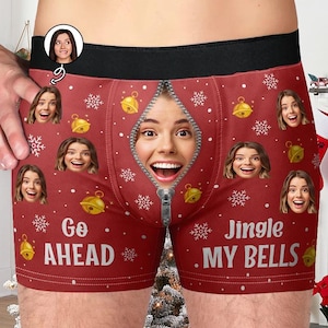 May include: Red boxer shorts with a black waistband, featuring a zipper design with a woman's face. The shorts are decorated with gold bells, snowflakes, and the phrases "Go Ahead" and "Jingle My Bells" in white text.