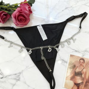 May include: Black thong underwear with a silver chain spelling "Daddy". The chain features small, sparkling details. The underwear is presented on a marble surface, with pink roses in the background.