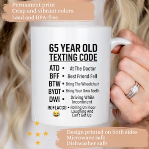 May include: A white ceramic mug featuring black text titled 65 YEAR OLD TEXTING CODE, listing humorous definitions for acronyms like ATD, BFF, BTW, BYOT, DWI, and ROFLACGU, accompanied by a laughing emoji. The mug has a handle on the right side.