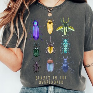 Könnte beinhalten: Ein anthrazitfarbenes T-Shirt mit einem Grafikdruck von neun bunten Käfern, die in einem Raster angeordnet sind. Unter den Insekten steht der Text "BEAUTY IN THE OVERLOOKED" in einer einfachen, hellen Schrift. Das Shirt hat einen Rundhalsausschnitt.