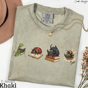 May include: A khaki-colored crew neck t-shirt featuring a graphic print of four insects reading books: a green praying mantis, a red ladybug, a dark beetle on a stack of books, and a bumblebee perched on a flower, all reading small books.