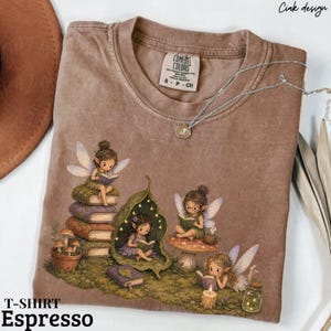 Può includere: Una t-shirt marrone a maniche corte con una stampa grafica di quattro fate che leggono libri in un ambiente boschivo. Le fate sono circondate da pile di libri, un fungo, un rifugio a forma di foglia e una candela. La maglia ha un girocollo.