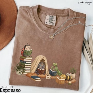 以下が含まれることがあります： 本を読む様々な昆虫のグラフィックが描かれた茶色のクルーネックTシャツ。イラストには、本の山に乗ったテントウムシ、テントの中にいるカブトムシ、カマキリ、ハチが描かれており、すべて本を読んでいる。Comfort Colorsのラベル付き。