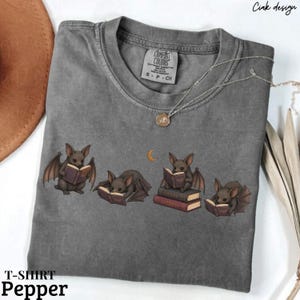 Peut inclure: Un t-shirt gris anthracite à manches courtes orné d'un motif représentant quatre petites chauves-souris brunes lisant des livres. Les chauves-souris sont illustrées dans diverses positions avec des livres ouverts et un petit croissant de lune jaune.
