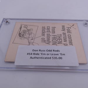 Puede incluir: Una tarjeta blanca con texto negro en un marco de plástico transparente. El texto dice: "Dobla la esquina, apunta la flecha hacia adelante, luego saca la tarjeta lentamente, quita la pegatina. Don Russ Odd Rods #54 Ride 'Em or Leave 'Em Autenticado 35,00 €"