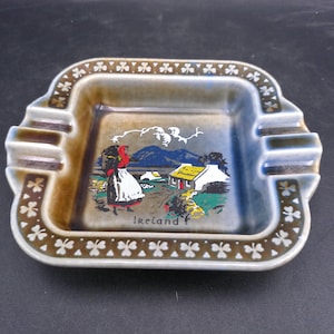 May include: Square ceramic ashtray with a brown and green shamrock border. The center features a colorful illustration of a woman in traditional Irish clothing walking past a cottage with the word "Ireland" printed below.